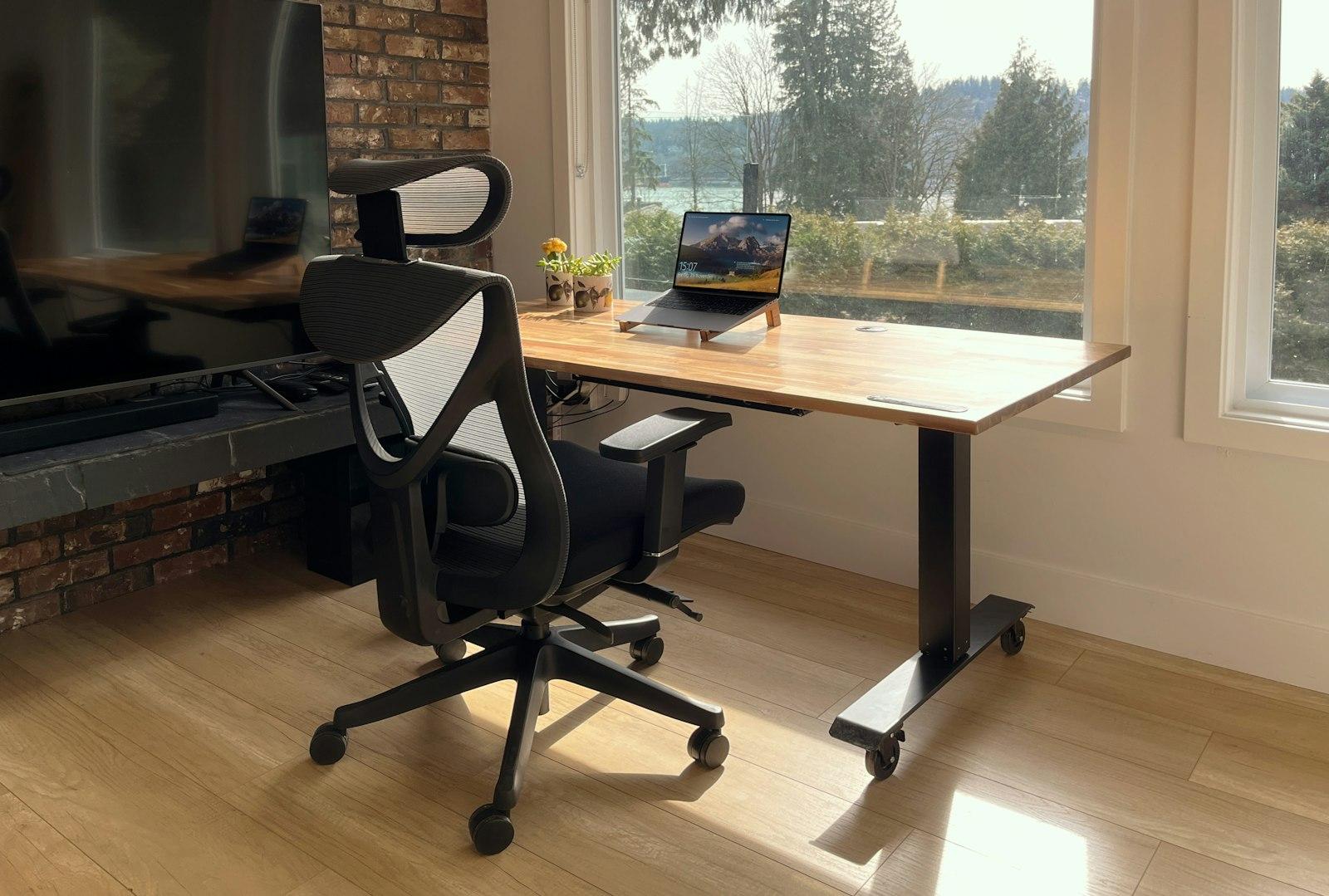 Bureau assis-debout au plateau bois clair devant une grande baie vitrée, avec chaise ergonomique à maillage
